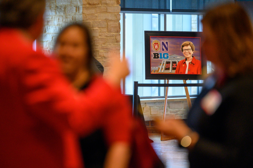 Looking across a large reception room, the photo focuses on an easel holding a photo collage of Rebecca Blank with Bascom Hall in the background as well as the UW–Madison crest, the Northwestern University N logo and the Big Ten logo. People attending the reception in the foreground of the photo are out of focus, centering attention on the portrait of Blank.