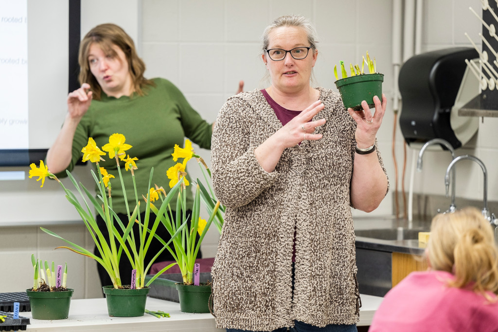 A woman stands at the front of a class and holds a green plastic plant container with multiple sprouting daffodils emerging from its soil. Behind her, another woman interprets her words into American Sign Language. Several more containers with daffodils in various stages of growth and blooming sit on a long table standing between the two women.