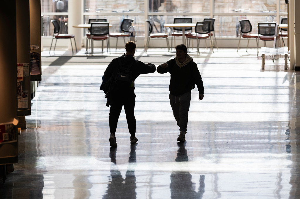 In a large corridor lit by floor-to ceiling windows at one end, two men appear in silhouette as the walk past each other in the hall. They reach out in greeting with an elbow bump.