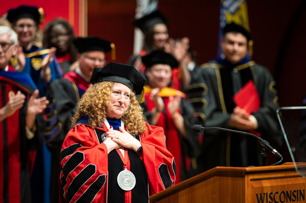 Jennifer Mnookin stands on stage with a crowd of dignitaries, all wearing academic regalia. Mnookin folds her hands on her heart in a gesture of gratitude as she makes an address from a wooden podium that has the word 