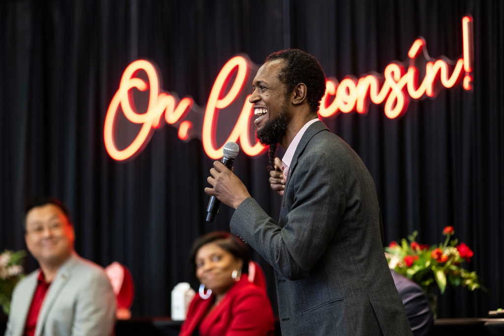 Chris Walker smiles as he stands and speaks into a microphone during a panel discussion. He's standing in profile to the camera and speaking to an audience that is out of frame. Behind him, hanging on the wall, a large neon sign reads, On, Wisconsin!