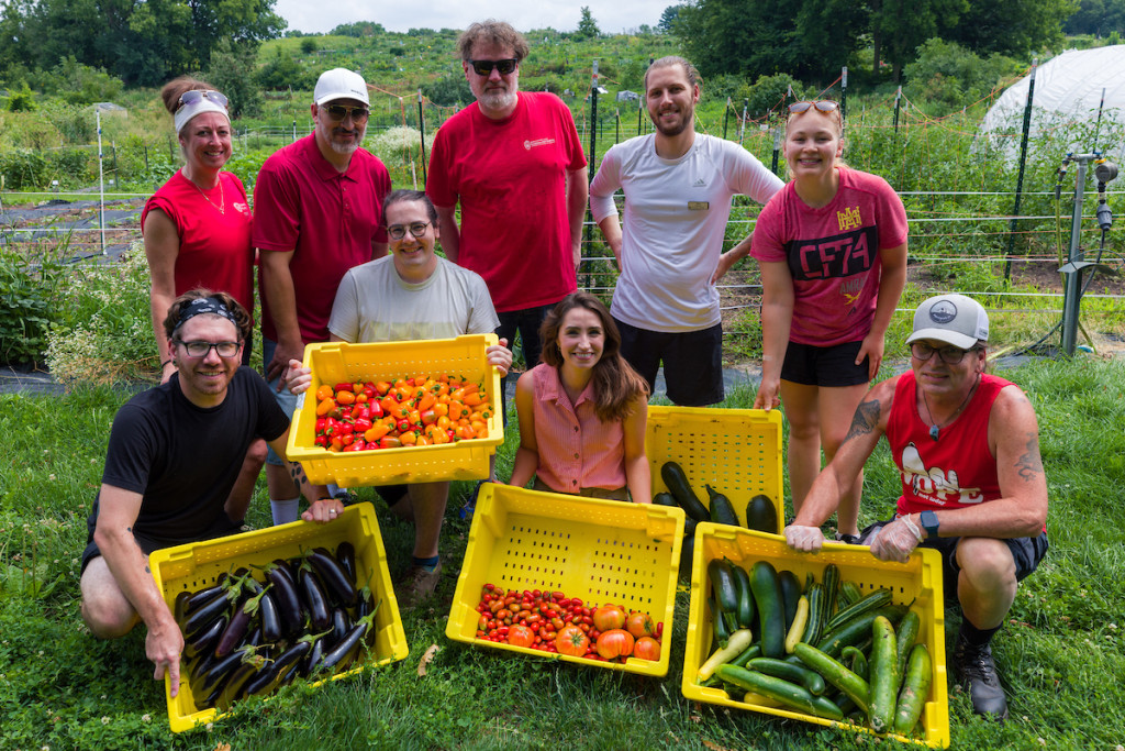 Eight people stand and display vegetables they picked, collected in large yellow bins.