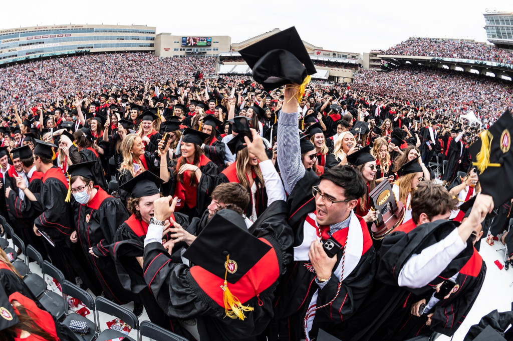 A wide-angle photo of Camp Randall Stadium taken during the spring commencement ceremony shows a sea of students wearing academic regalia. They're laughing, jumping and celebrating.