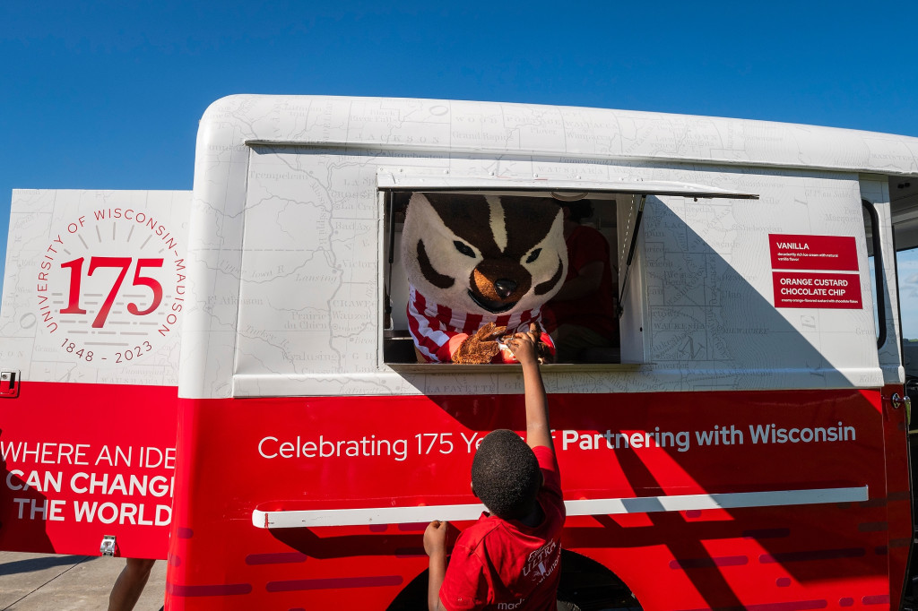 Bucky Badger leans out of a service window on the side of a vintage dairy truck that's been decorated in red and white for UW's 175th anniversary. He's handing an ice cream cup to a child who's reaching up to accept it.
