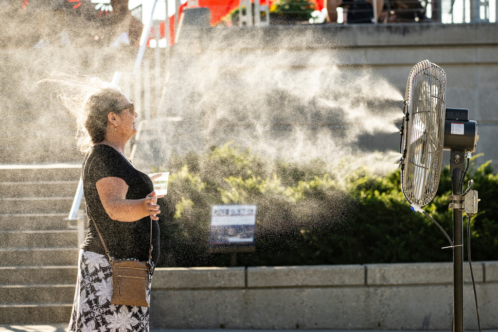 A woman stands in profile to the camera with her arms outstretched and her eyes closed behind her sunglasses. She's standing outside on the Memorial Union Terrace while a fan blows a mist of cool water over her face and arms.