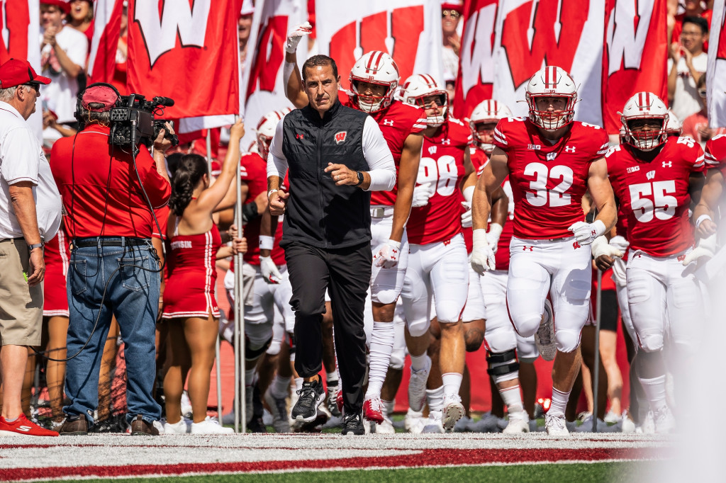 Amid red and white flags bearing the letter W, football coach Luke Fickell runs onto the football field, followed by football players wearing red and white jerseys.