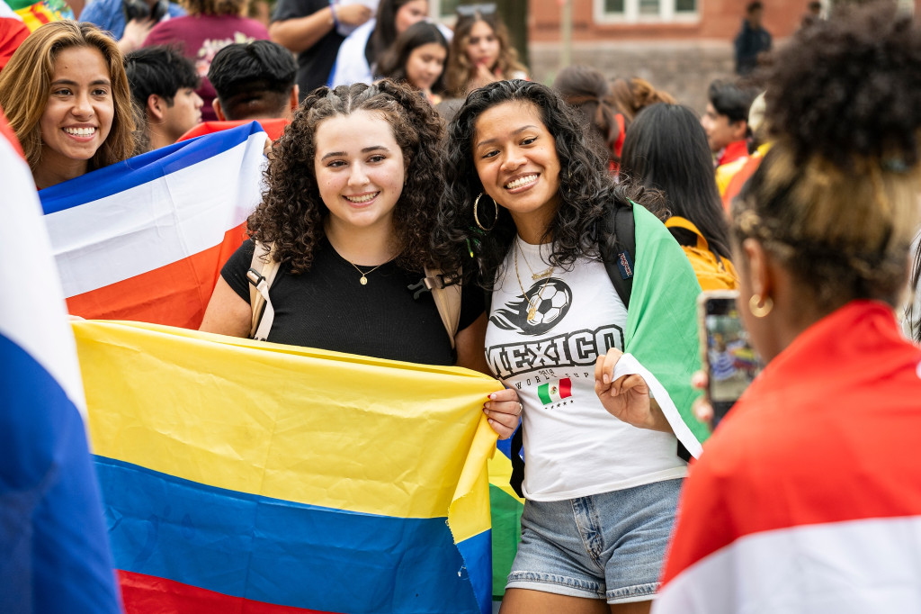 Three women stand together in a crowd on Bascom Hill. They're smiling for a photo while holding up the flags of Chile, Colombia and Mexico.