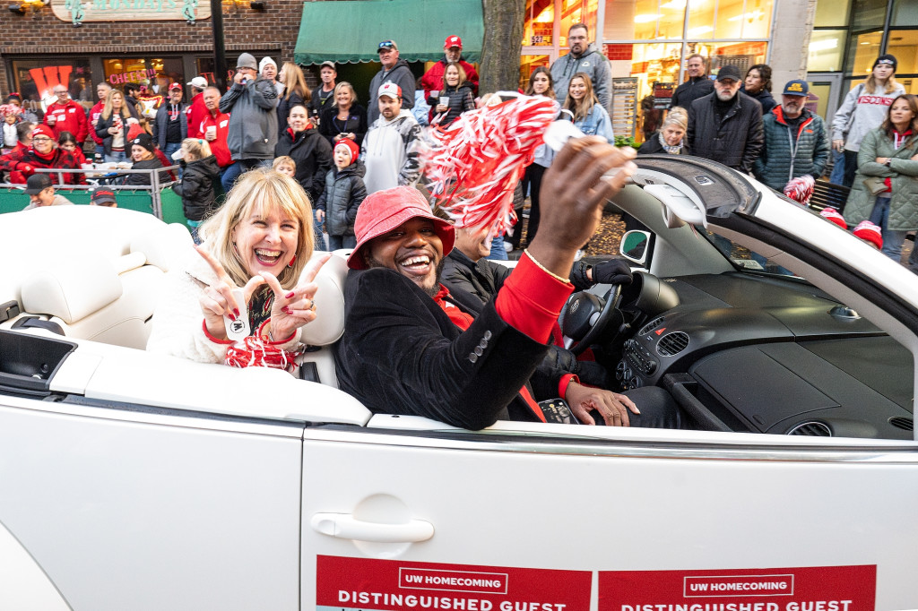 Lori Reesor and LaVar Charleston ride down State Street during the homecoming parade in a white VW Bug convertible. Reesor is making the W sign with her thumbs and forefingers while Charleston is waving a red and white pom-pom. They're both smiling to the camera while the crowd looks on.