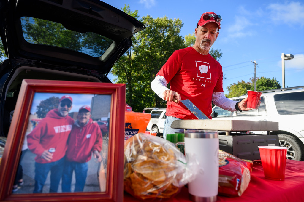 A man waring a red and white UW–Madison hat and T-shirt flips burgers on a griddle set up on a tabletop in a parking lot near Camp Randall stadium. In front of him on the table is a photo of himself with his father during a previous year's homecoming tailgate.