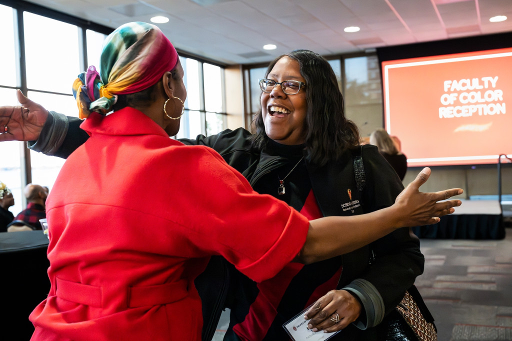 In a reception room, two women, Angela Byars-Winston and Linda Scott smile and move to embrace each other. Behind them, a screen at the front of the room reads Faculty of Color Reception.