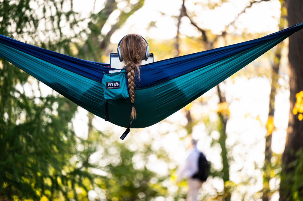 A student's long braid falls out of the back of a hammock hung between two trees on UW's Lakeshore Path. The student has headphones on and is looking at a laptop screen.