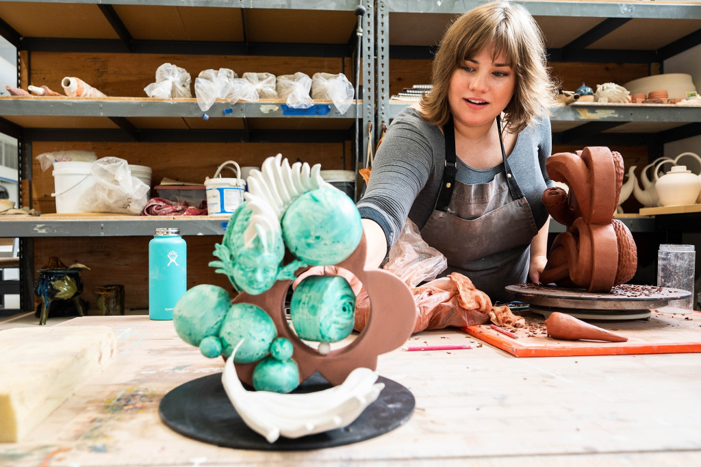 A woman leans over a work table in a ceramics studio to make an adjustment to a sculpture sitting on the table.