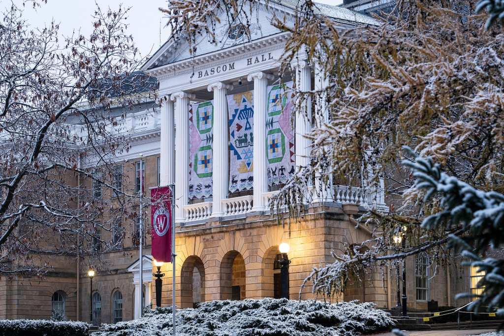 A view of the front of Bascom Hall through snow-covered branches. The Seed by Seed banner hangs between tall, white columns above the building's main entrance. The banner has been printed with a texture resembling beadwork and contains symbols and colors representing traditions of the Ho-Chunk Nation. Four green rings represent the four lakes of Teejop, the name the Ho-Chunk give the land now occupied by UW–Madison. Inside each ring, square patches in light blue, dark blue yellow and red represent the reflections of light on the water at different times of day. Two large pink triangles on either side of the banner represent flowers, with green stems and triangular leaves leading to the center panel. On the center panel, a large diamond made of small blue triangles frames a blue thunderbird, which is flanked by two red, abstract W's, representing UW–Madison. Below the thunderbird are two green water spirits, which resemble four-legged animals with very long tails. Below the water spirits are six light blue triangles representing water. Above and below the large diamond frame are bursts of yellow beading, representing the sun. Along the bottom border of the banners are stylized animal symbols of the twelve clans of the Ho-Chunk Nation, and beneath each animal is a traditional Ho-Chunk flower motif in blue and green.