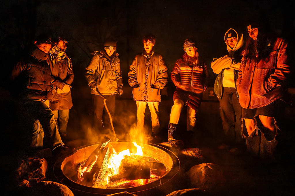 In a photo lit only by the light of a bonfire, a group of seven students dressed in heavy coats and hats gather around the flames and share stories.