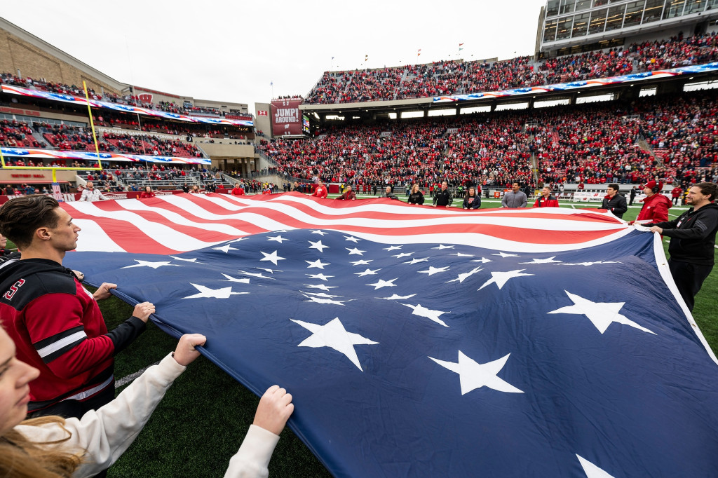 Fans fill the stands at Camp Randall while a team of people hold the edges of a giant U.S. flag on the field.
