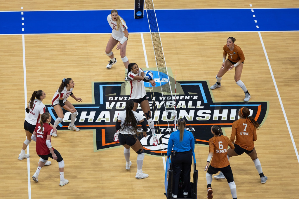 The UW–Madison volleyball team faces off against the University of Texas. One player on the UW side bumps the ball while another moves it to set it. On the court floor are the words, NCAA Division I Women's Volleyball Championship 2023 Tampa.