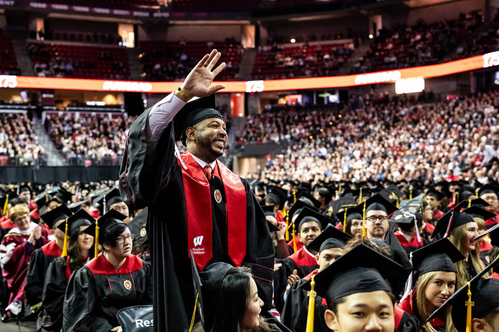 Wearing academic regalia, Devin Harris stands among a crowd of seated graduates at commencement. He smiles and waves with one hand.