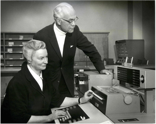 In a black and white photo of a mid-twentieth-century computer lab, a woman sits at a computer control deck while a man stands behind her to learn what she is doing.