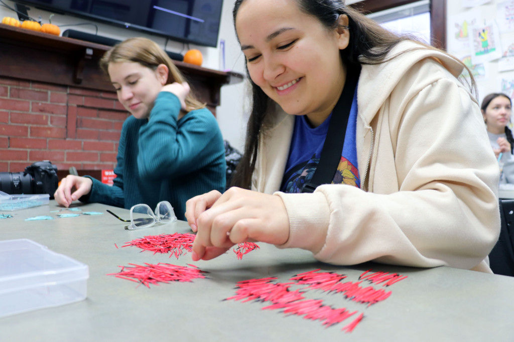 A young woman sits at a table smiling while organizing red-dyed quills for a craft project.