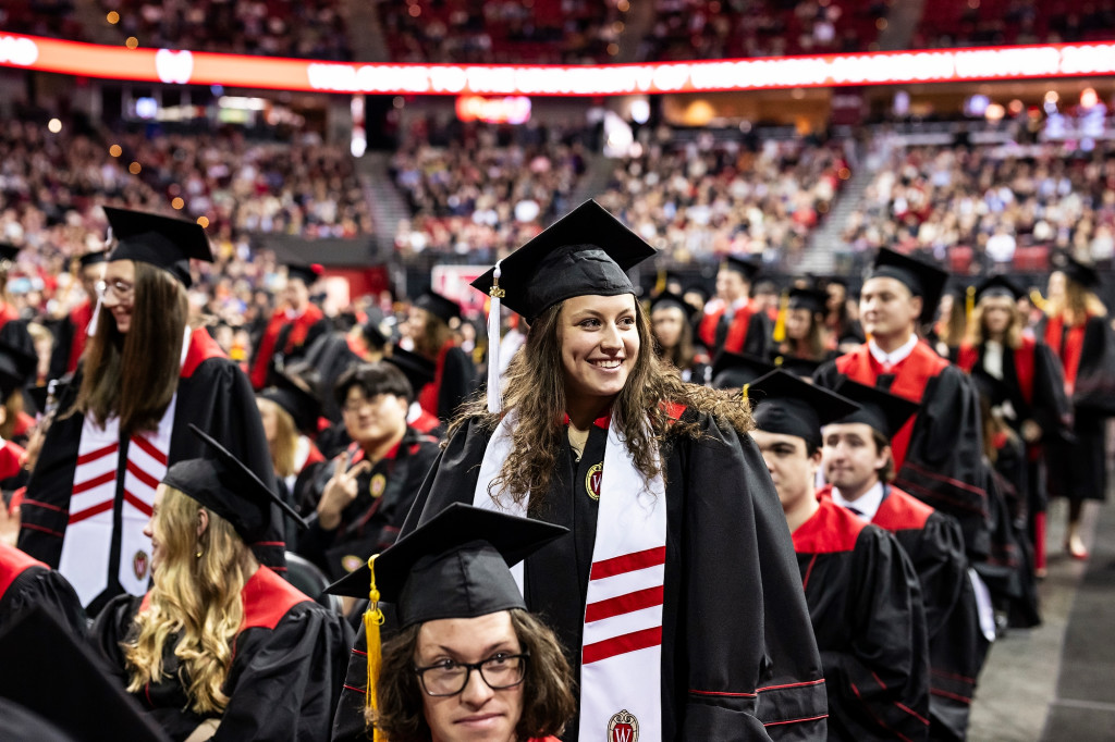 A woman wearing a white-and-red stole stands and smiles amid the graduates in the Kohl Center.