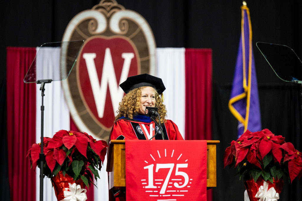 A woman wearing academic robes stands at a podium and speaks with a W crest logo behind her.