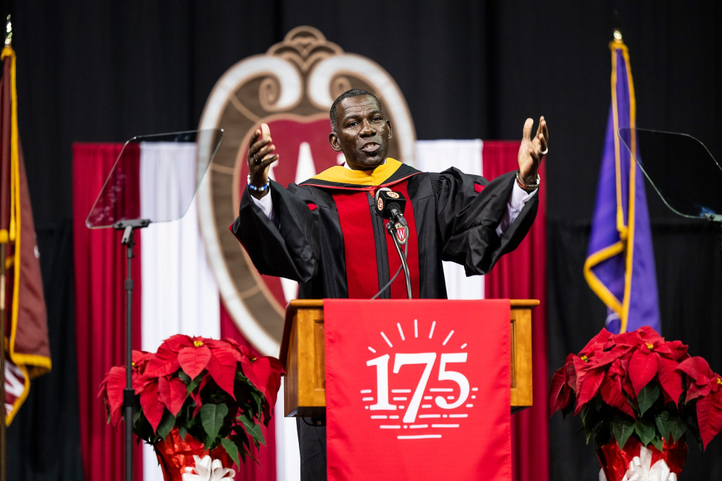 A man wearing an academic robe gestures as he speaks at the podium.