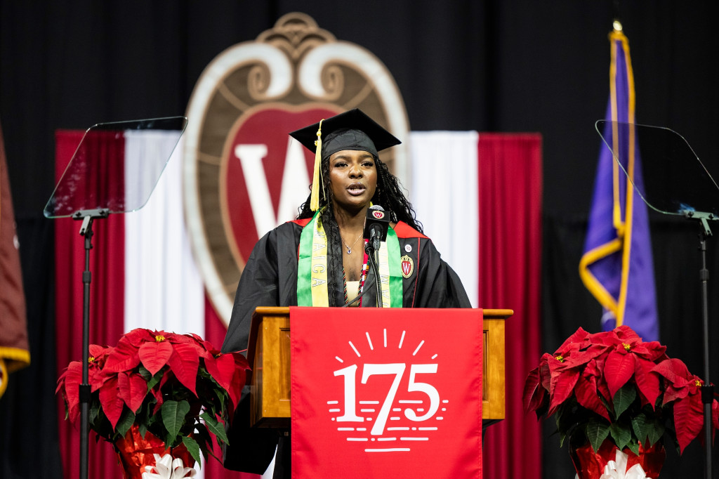 A woman stands at a podium and speaks; she wears commencement finery.