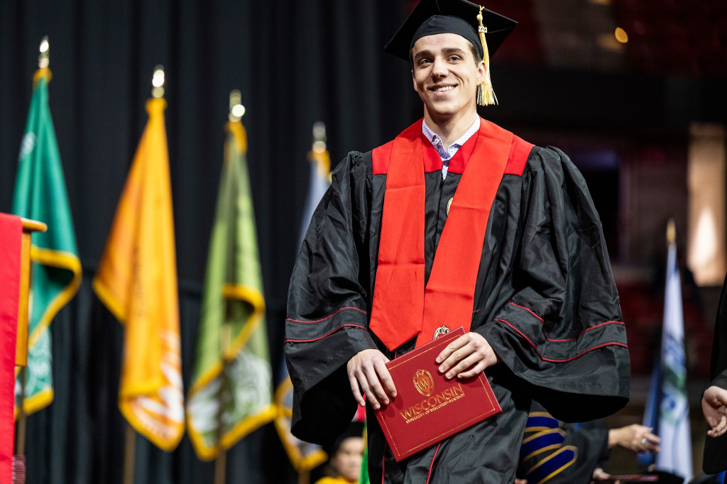 A graduate with a black gown and red stole walks across a stage; flags are visible in the background.