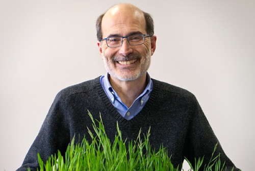 A studio photo, a smiling man from the waist up. He's standing behind a tray of green, leafy grass, which spans the bottom of the frame.
