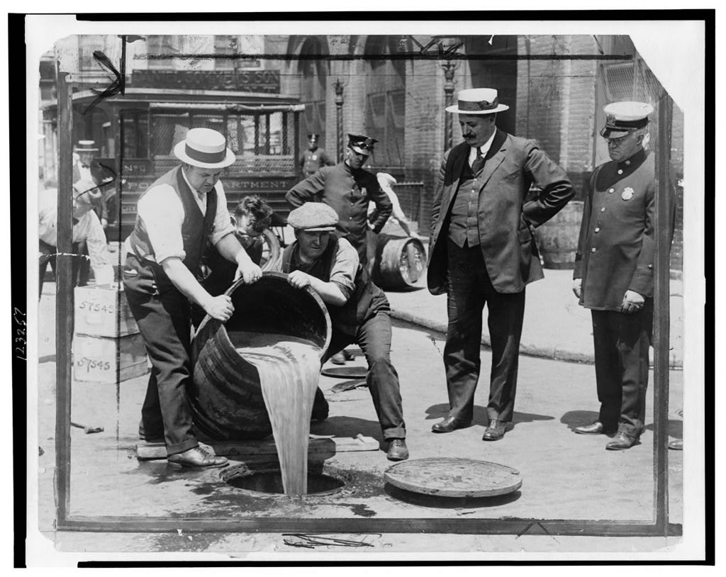 In an old black and white photo from the 1920s, a man in a suit and hat stands next to a police officer as they watch two other men pour a wooden barrel full of beer into a sewer drain in the middle of the street.