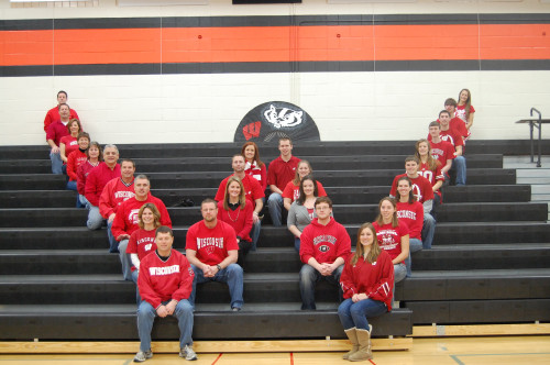 Photo of 27 people sitting in a letter W formation on bleachers in a high school gym. They're all wearing blue jeans and red and white UW–Madison shirts.