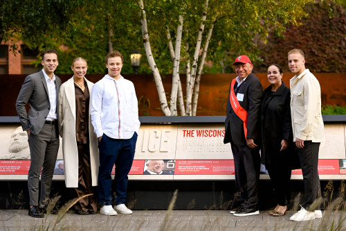 A photo of six people standing outdoors in front of a display in Alumni Park at UW–Madison. They're smiling at the camera as they stand on either side of a placard honoring Elzie Higginbottom, fourth from left and wearing a red sash.