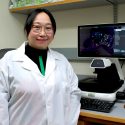 A woman in a lab coat stands in front of a computer screen.