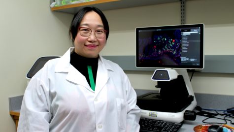 A woman in a lab coat stands in front of a computer screen.