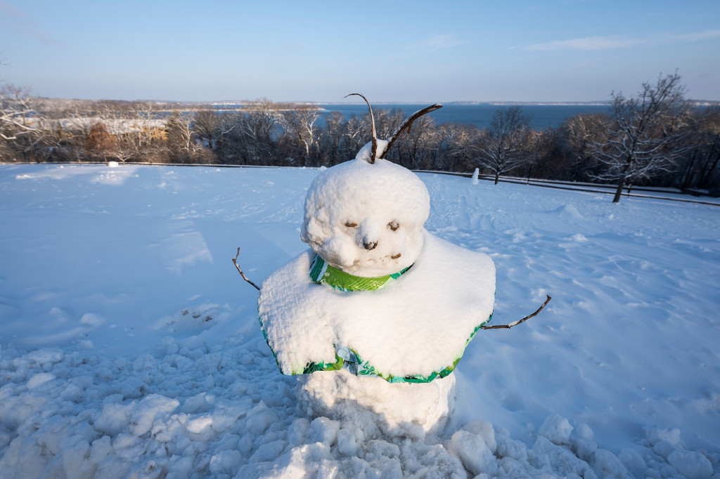 The sun shines upon a snowman positioned atop snow-covered Observatory Hill at the University of Wisconsin–Madison during winter on Jan. 11, 2024. In the background is the still open and unfrozen water of Lake Mendota.