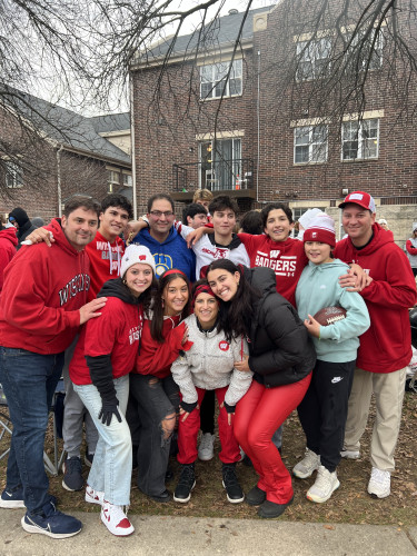 Standing outside a brick apartment building on a cloudy October day, a group of 11 members of the Rivera family bunch together in two rows to smile for a photo. They're wearing red and white UW–Madison shirts, hats and hoodies.