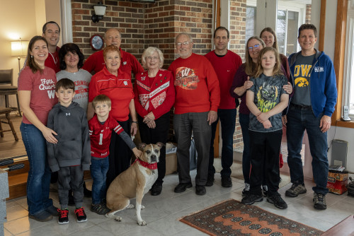 Fourteen members of the Schellin family and the family dog stand together and smile to the camera. They're wearing red and white Badger gear. The dog is wearing a red bandanna.