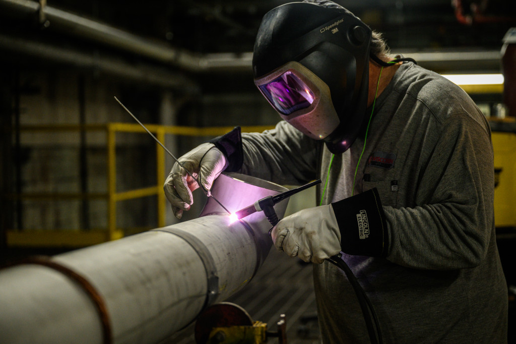 A man uses a piece of equipment to weld, the flame lighting up a dark room.