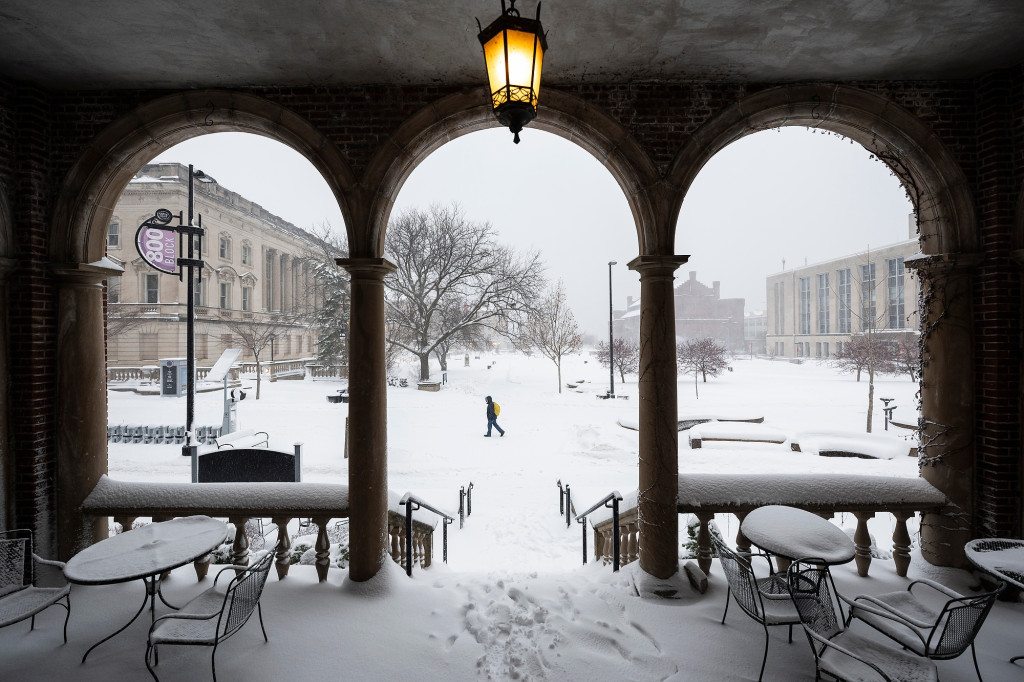 A person walks along snowy campus sidewalks; in the foreground are building arches.