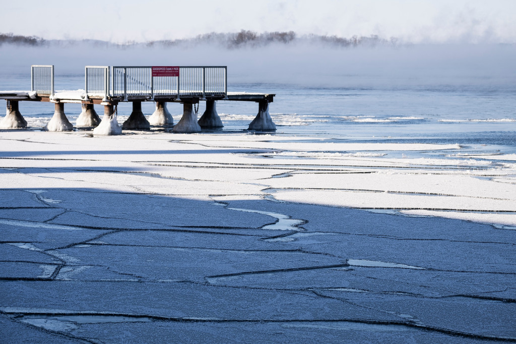 Ice starts to form, with a thin coat of snow on it, on a lake. A dock is in the background.