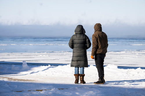 Two people stand on the shore next to a frozen lake, looking out.