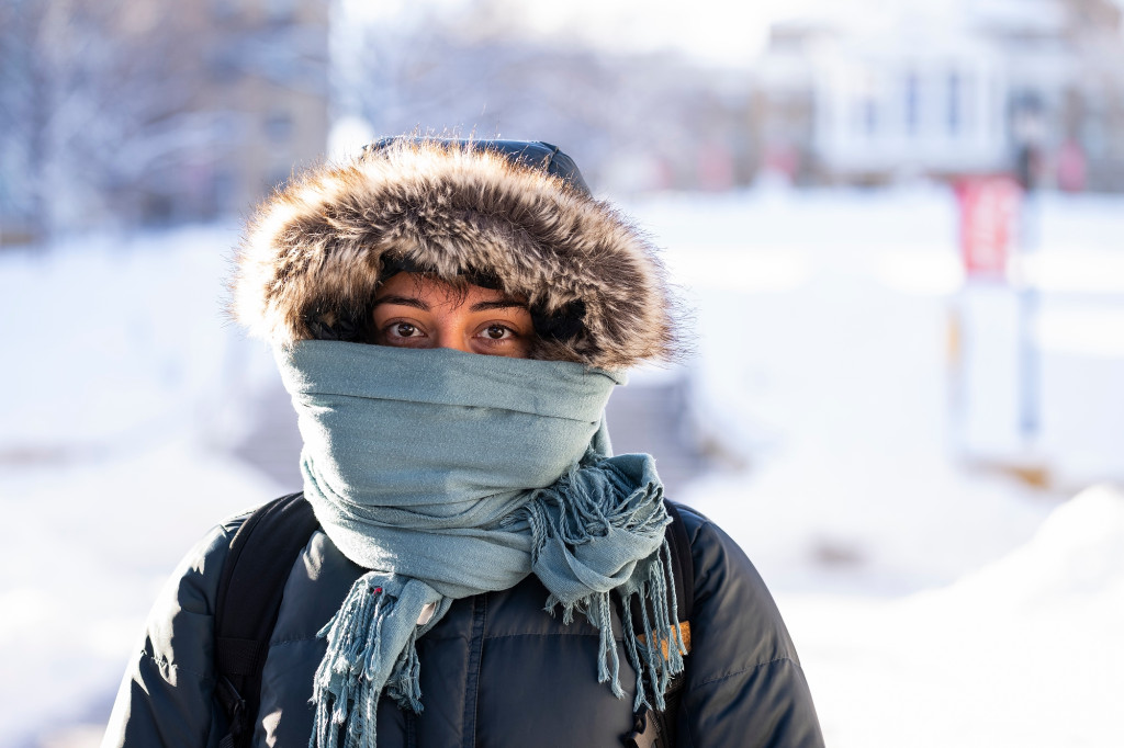 A woman wearing a parka and with a scarf draped over her face looks at the camera.
