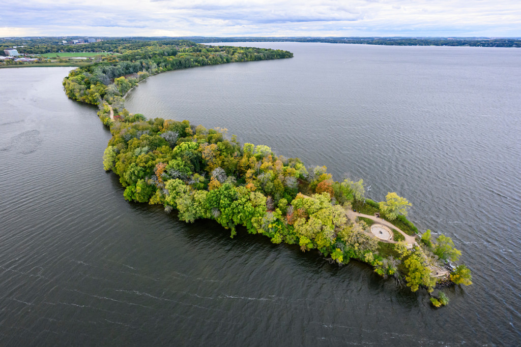 An aerial photo of green, tree-covered Picnic Point, a narrow peninsula stretching into Lake Mendota.