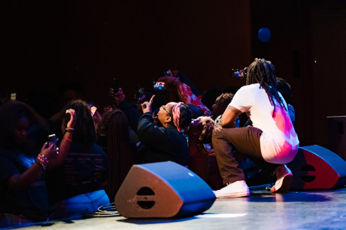 A visiting musical artists crouches down while performing on stage to take selfie photos with the audience.