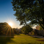 Photo of a sunrise on campus, with rays of sun streaming over green lawns and historic buildings.