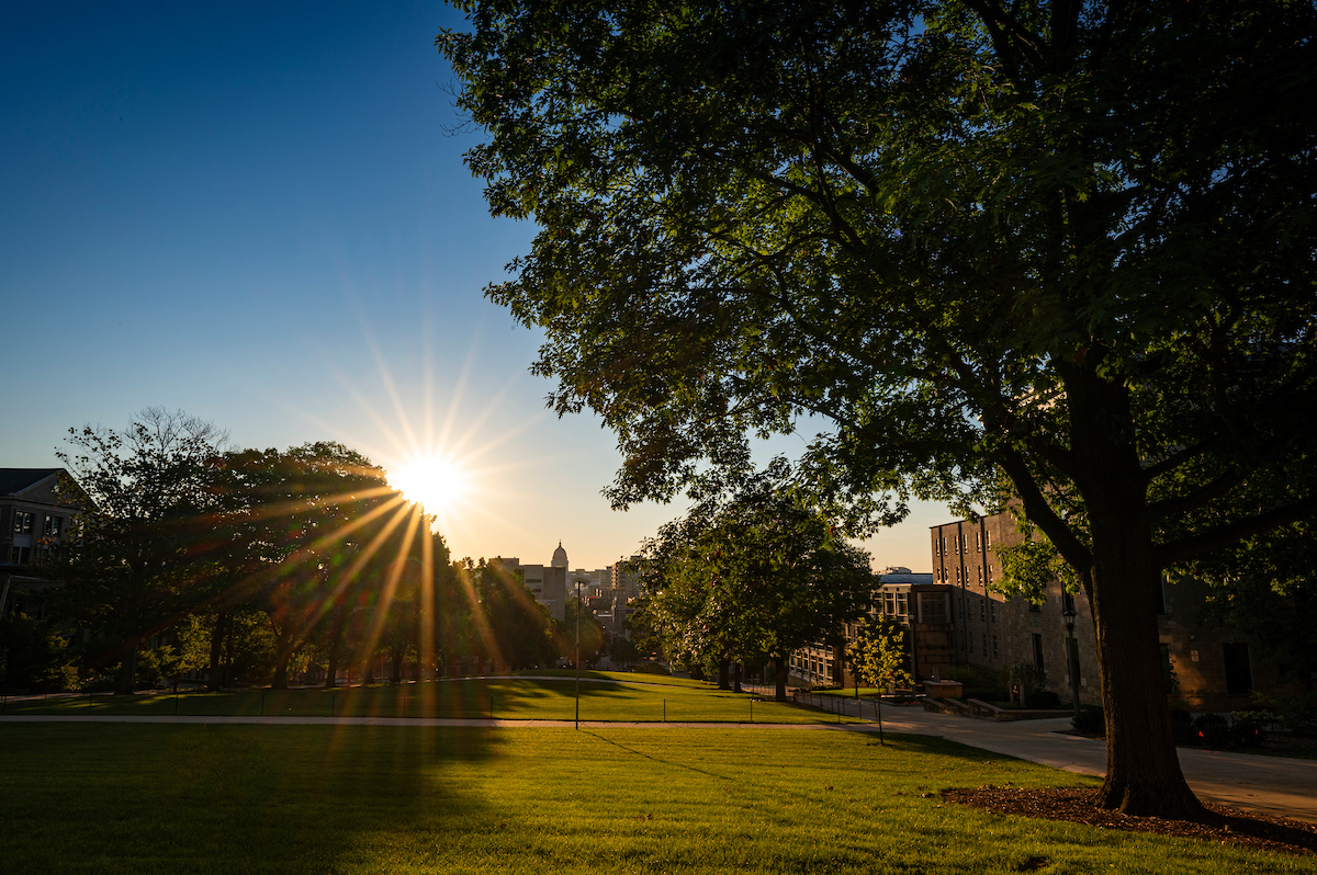 Photo of a sunrise on campus, with rays of sun streaming over green lawns and historic buildings.