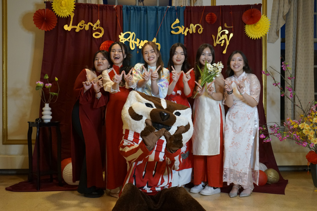 A photo of six women wearing traditional Vietnamese dress stand in a photo booth with Bucky Badger. They're all making W signs with their hands.