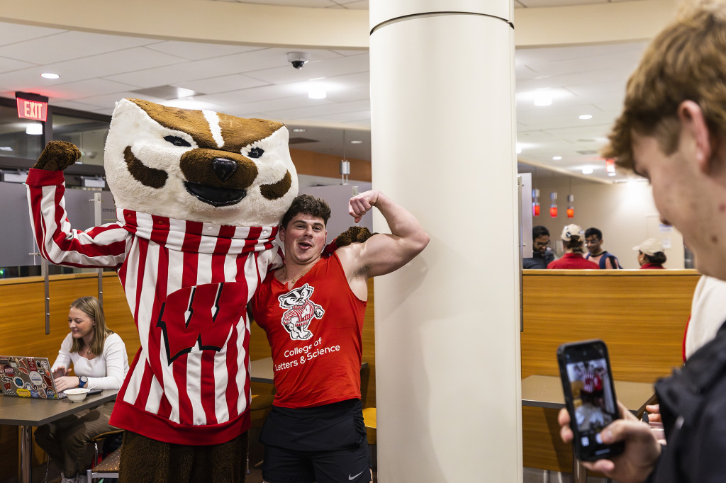 A young man and Bucky Badger, arm in arm, stand looking at the camera and flexing their biceps.