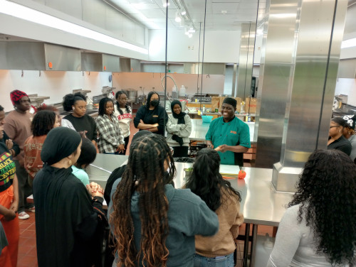 Students stand gather in a demonstration kitchen listening to a local chef talk about food.