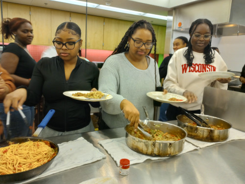 Students stand in a line behind a table dishing up the food they recently prepared.